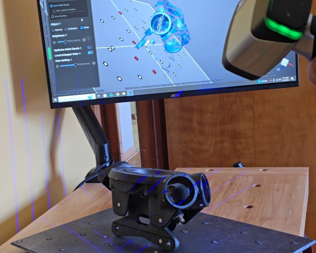 A 3D laser scanner handheld in the upper right, positioned over a monitor displaying a blue 3D model and a black laser scanner mounted on a metal plate on a wooden table. Purple laser lines are visible on the table.